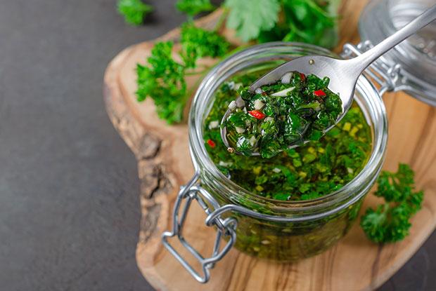 Un pot en verre avec une sauce chimichurri fraîche faite maison, posé sur une planche en bois avec des herbes sur fond vert